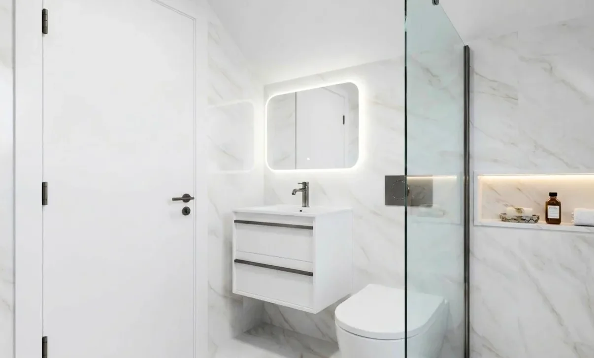 DDL design visual of the Aldenham guest bathroom: a white marble-look tile with soft grey and gold veining on the walls and floor, a wall-hung white vanity under a backlit mirror, a glass shower screen and a lit niche set into the shower wall